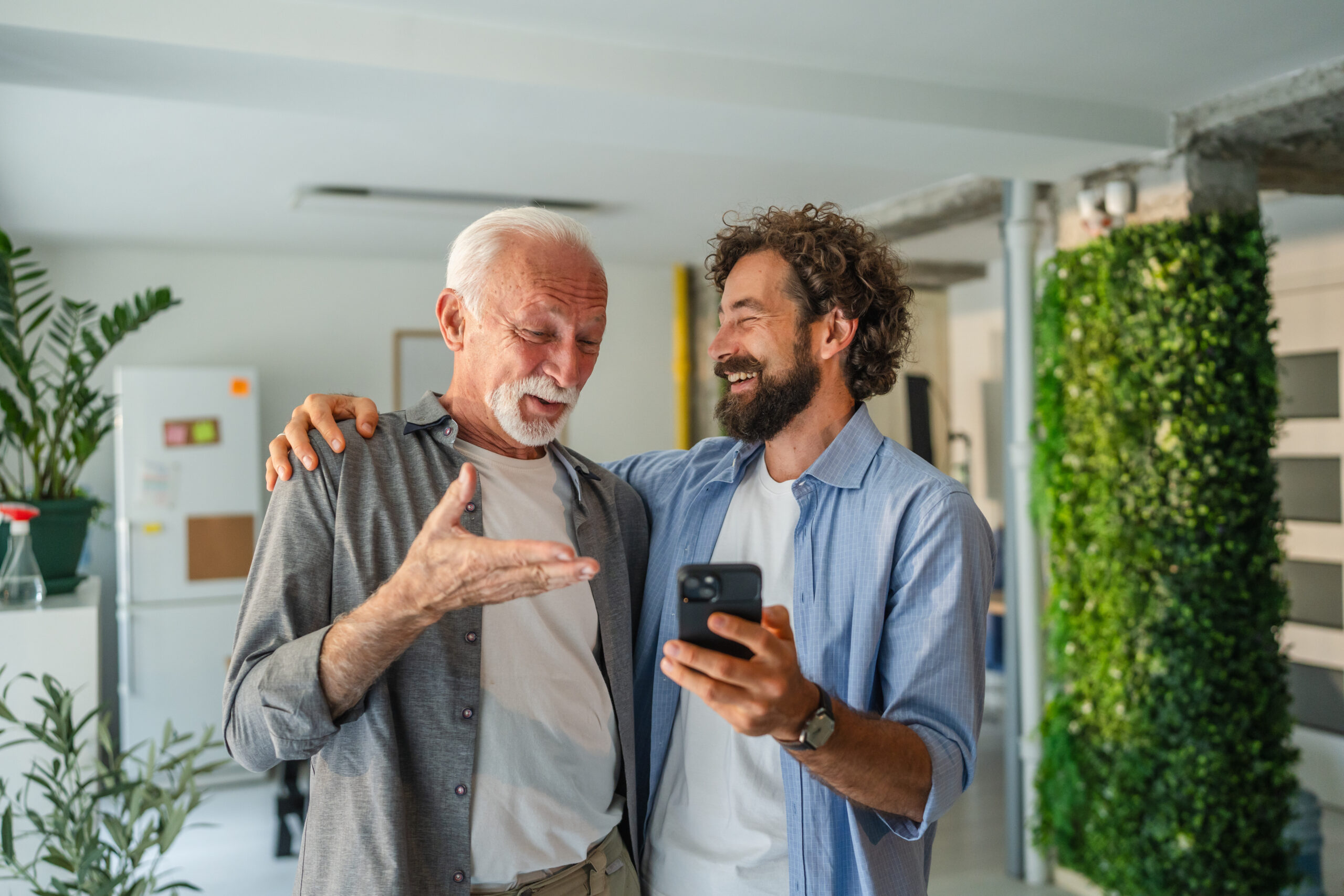 Adult son smiling while showing senior father how to use a smart phone, pointing at the screen in a bright, inviting home interior, fostering connection and support between generations