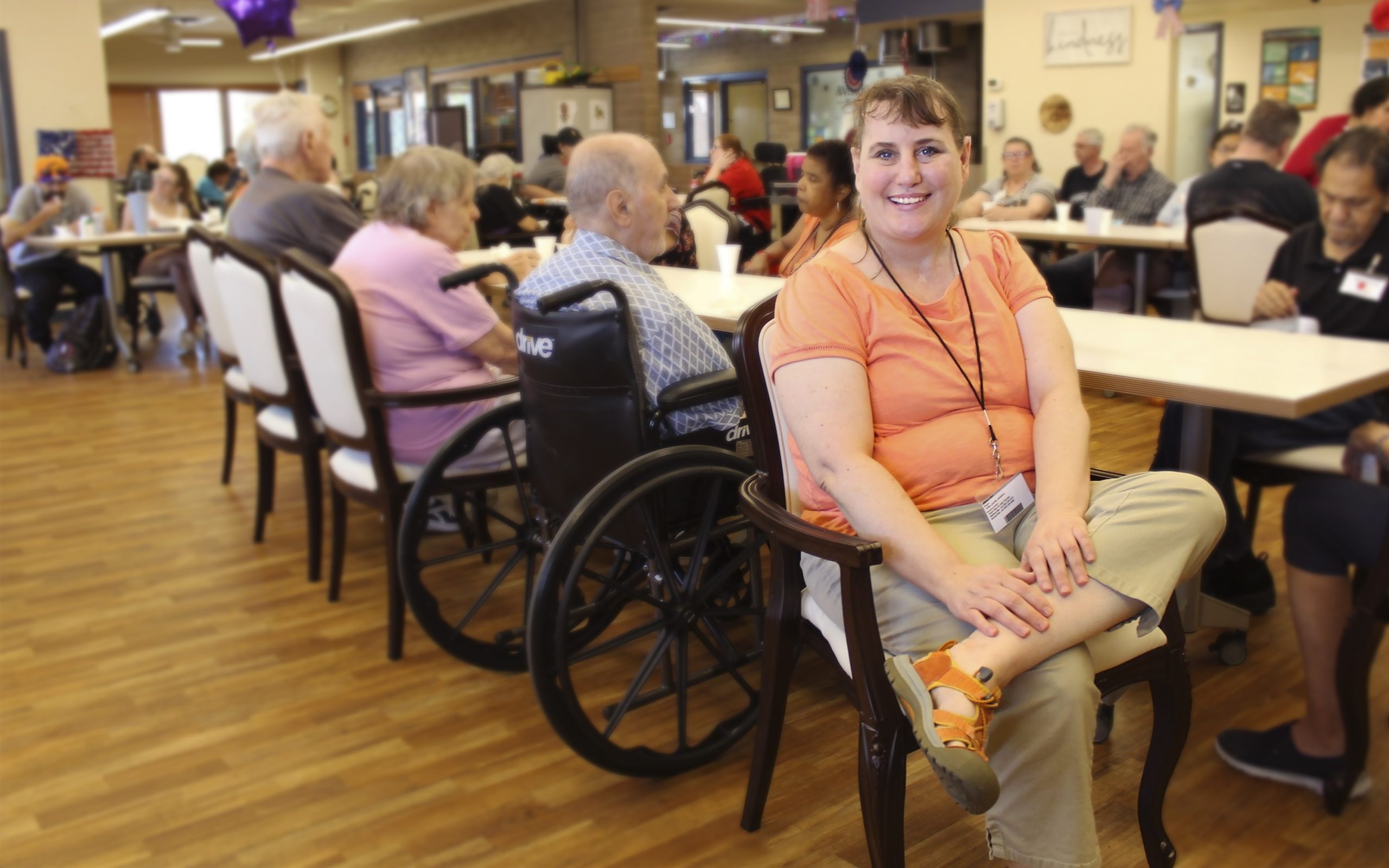 A woman sitting in her chair smiling at the camera with a group of senior behind him